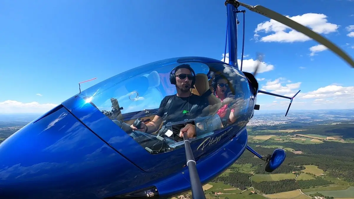 Pilote et passager casqués à bord de l'autogire au-dessus des champs et des forêts